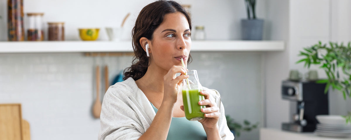 Woman wearing wireless earbuds drinking a green smoothie through a straw in a bright, modern kitchen.